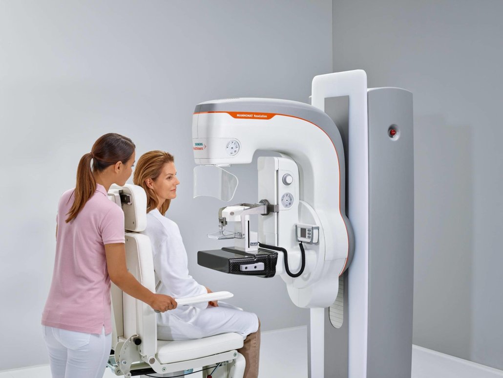Sitzende Aufnahme bei Tomosynthese während Mammographie Tomosynthese mit dem Siemens Healthineers Gerät Mammomat Revelation - Frau sitzt vor Geraet